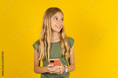 blonde kid girl wearing green T-shirt over yellow studio background hold telephone hands read good youth news look empty space advert