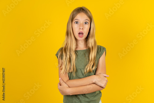 Fotografia Shocked embarrassed blonde kid girl wearing green T-shirt over yellow studio background keeps mouth widely opened