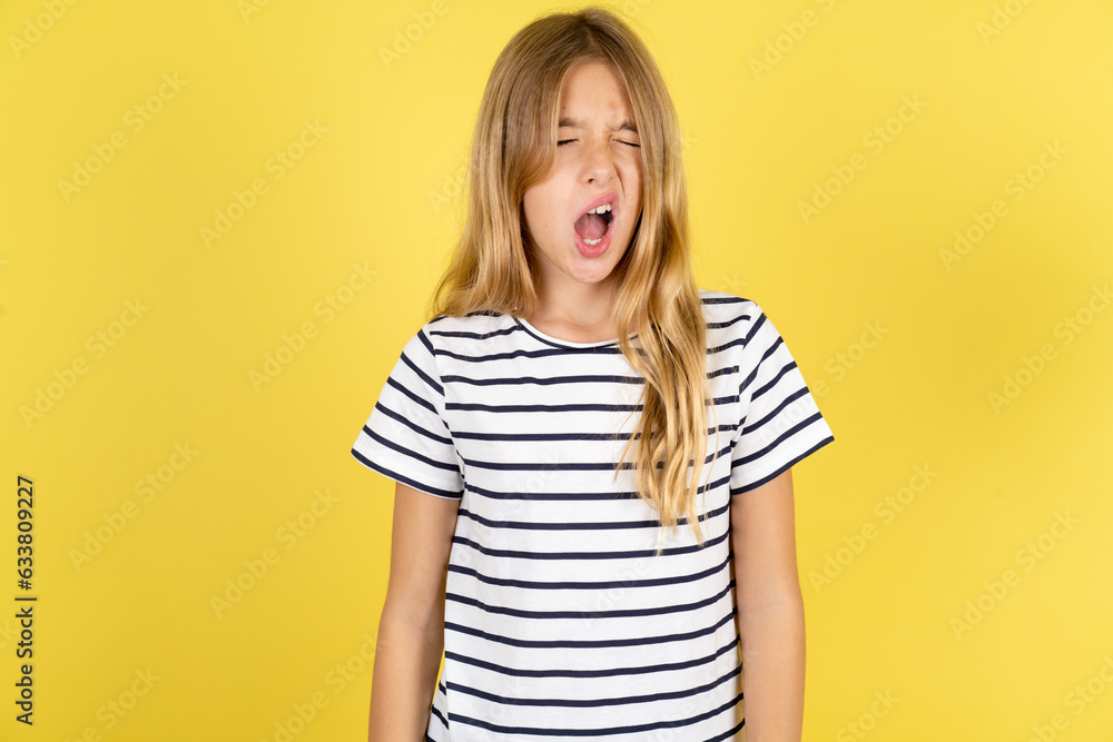 young beautiful blonde kid girl over yellow studio background yawns with opened mouth stands. Daily morning routine young beautiful blonde kid girl over yellow studio background yawns with opened mouth stands. Daily morning routine