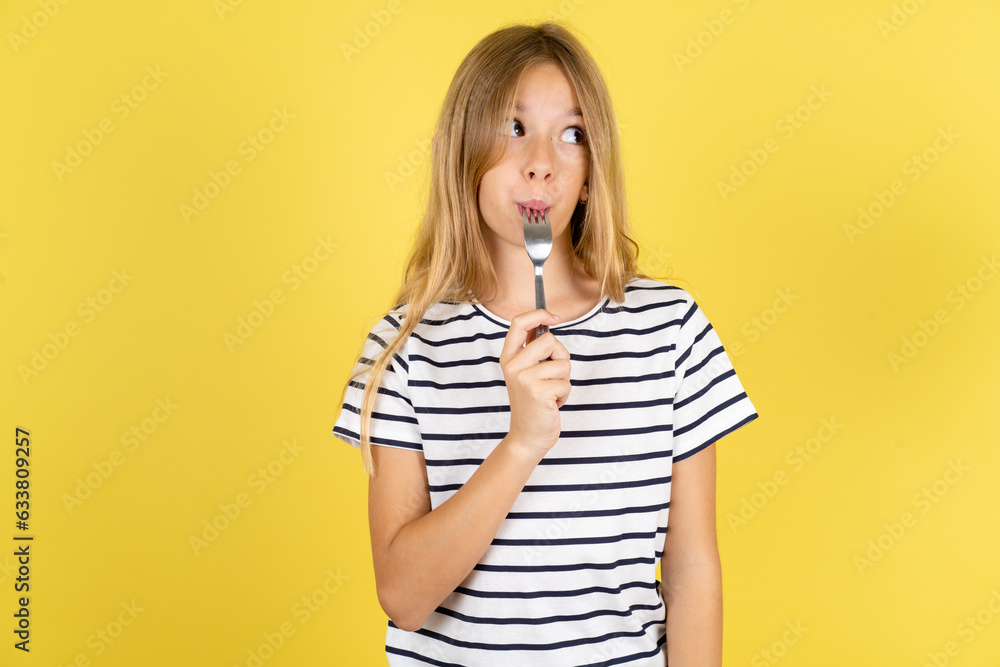 Photo of dreamy young beautiful blonde kid girl over yellow studio background lick fork look empty space Photo of dreamy young beautiful blonde kid girl over yellow studio background lick fork look empty space
