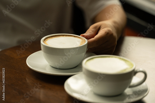 Closeup hand holding white cup of hot coffee latte and blurry hot green tea cup on wood background in restaurant.Best of menu in the coffee shop.Soft focus.