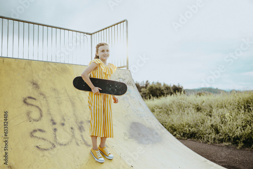 Mädchen, 9 Jahre, im gelbweiß gestreiften Jumpsuit mit Skateboard auf einer gelben Skaterbahn photo