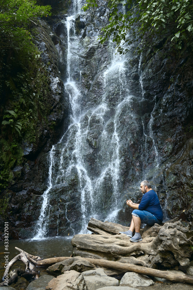 one, alone Asian man sitting and looking at screen of  mobile phone. smart phone.front view of Mirveti waterfall.