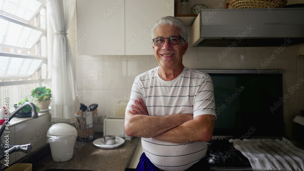Happy senior man standing in home kitchen by window with arms crossed smiling at camera Happy senior man standing in home kitchen by window with arms crossed smiling at camera