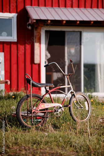 A vintage red and white bicycle with wide handlebars and a large seat propped up outside a red barn 