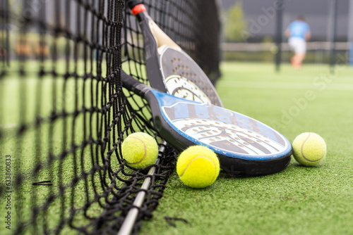 Yellow balls on grass turf near padel tennis racket behind net in green court outdoors with natural lighting. Paddle is a racquet game. Professional sport concept with copy space.