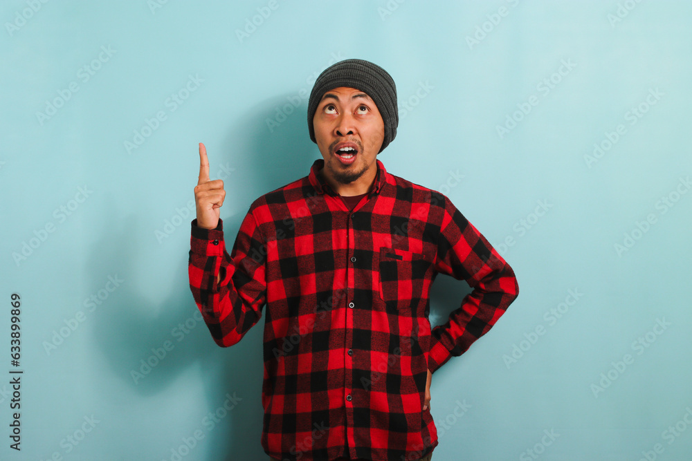 Surprised Young Asian man with a beanie hat and red plaid flannel shirt is pointing his finger up towards the copy space, isolated on a blue background Surprised Young Asian man with a beanie hat and red plaid flannel shirt is pointing his finger up towards the copy space, isolated on a blue background