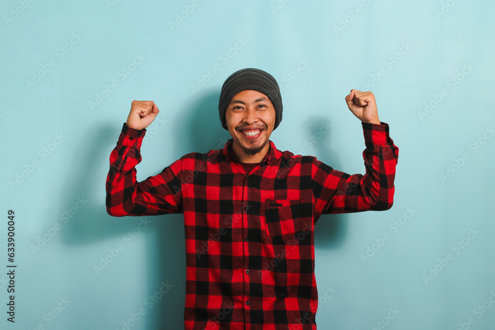 Enthusiastic young Asian man with beanie hat and red plaid flannel shirt rejoicing, saying yes, looking happy, and celebrating victory with a champion fist pump gesture, isolated on blue background Enthusiastic young Asian man with beanie hat and red plaid flannel shirt rejoicing, saying yes, looking happy, and celebrating victory with a champion fist pump gesture, isolated on blue background