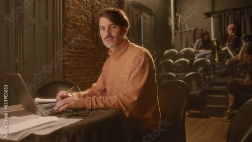 Professional male playwright sitting at table with laptop and papers and posing for the camera with smile, working on stage in a theater. Medium long shot, video portrait photo