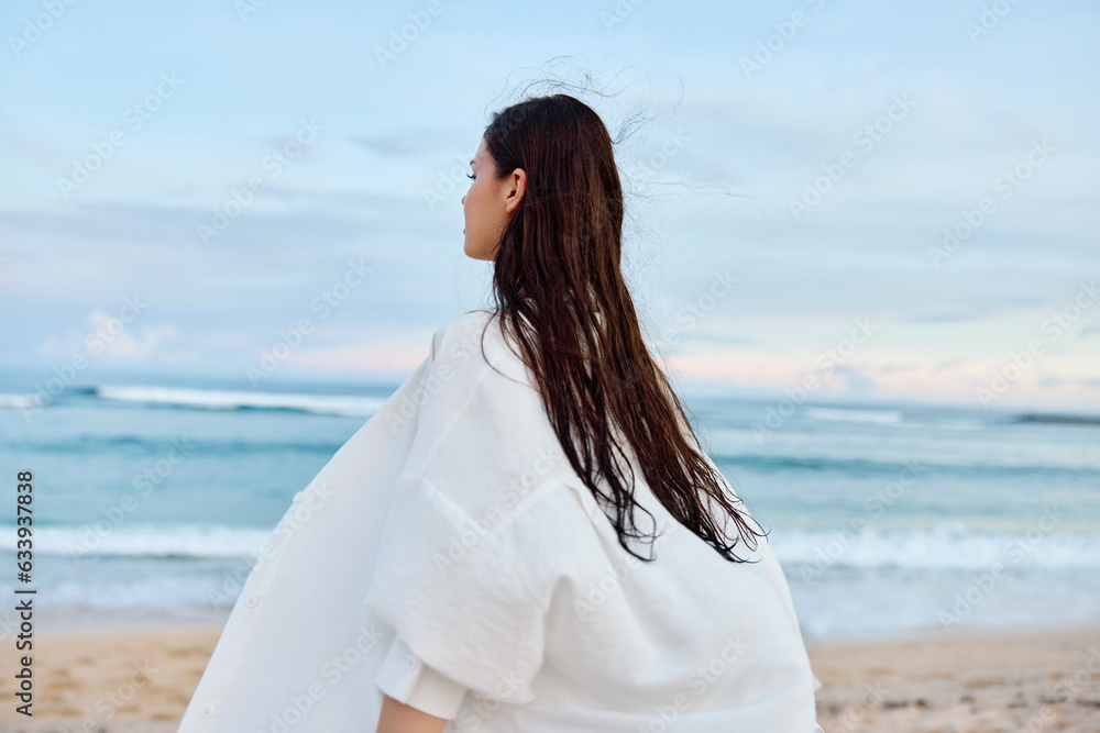 Happy tanned slender woman in white swimsuit shirt and denim shorts walks on the beach with her back to the camera on the sand by the ocean with wet hair after swimming, sunset light Happy tanned slender woman in white swimsuit shirt and denim shorts walks on the beach with her back to the camera on the sand by the ocean with wet hair after swimming, sunset light