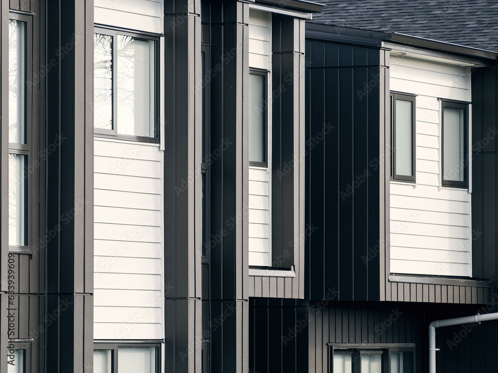 Close-up of row of attached modern newly built two storeys residential townhouses with windows. Horizontal white and vertical black weatherboards exterior walls design. Close-up of row of attached modern newly built two storeys residential townhouses with windows. Horizontal white and vertical black weatherboards exterior walls design.