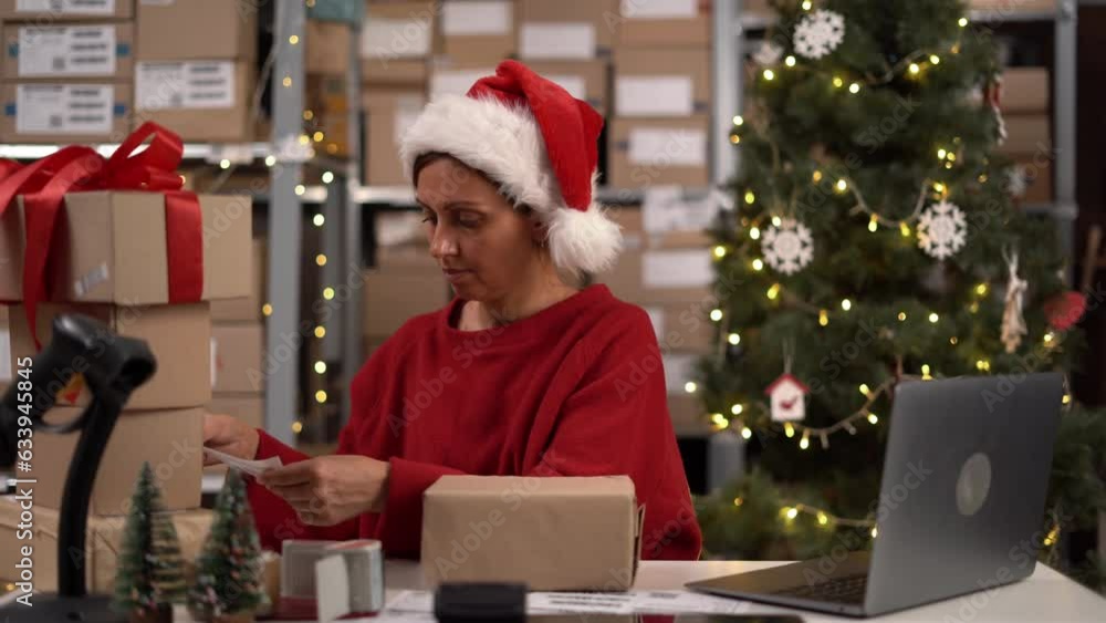 Delivery business. Woman in Santa hat with laptop applying the shipping label to parcels working in the warehouse or post office. Delivery business. Woman in Santa hat with laptop applying the shipping label to parcels working in the warehouse or post office.
