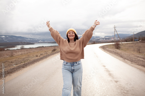 Beutiful woman is wearing jeans, sweater and hat walking in cold weather along the road        against the background of landscape and nature. A plus size girl is travelling