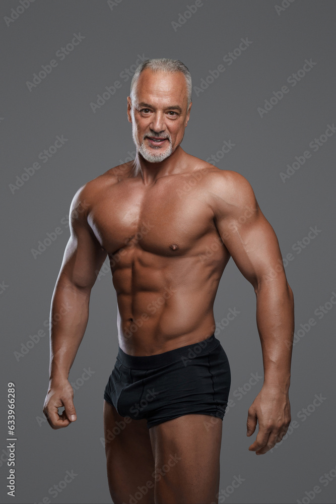 Attractive older male with muscular physique, stylish gray beard, black underwear and bare torso, posing against a gray backdrop
