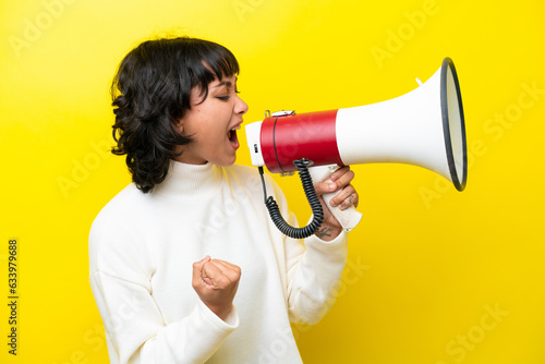 Young Argentinian woman isolated on yellow background shouting through a megaphone to announce something in lateral position