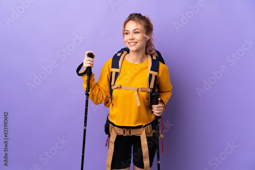 Teenager girl with backpack and trekking poles over isolated purple background posing with arms at hip and smiling