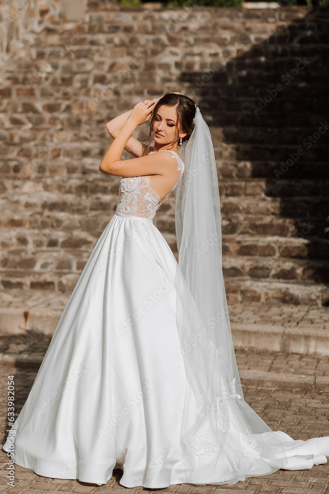 Portrait of a beautiful bride in a white satin voluminous dress with a long veil. A brunette girl with a beautiful hairstyle and professional make-up.