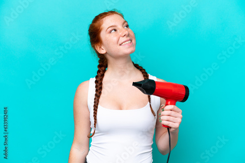 Young caucasian reddish holding a hairdryer isolated on blue background looking up while smiling
