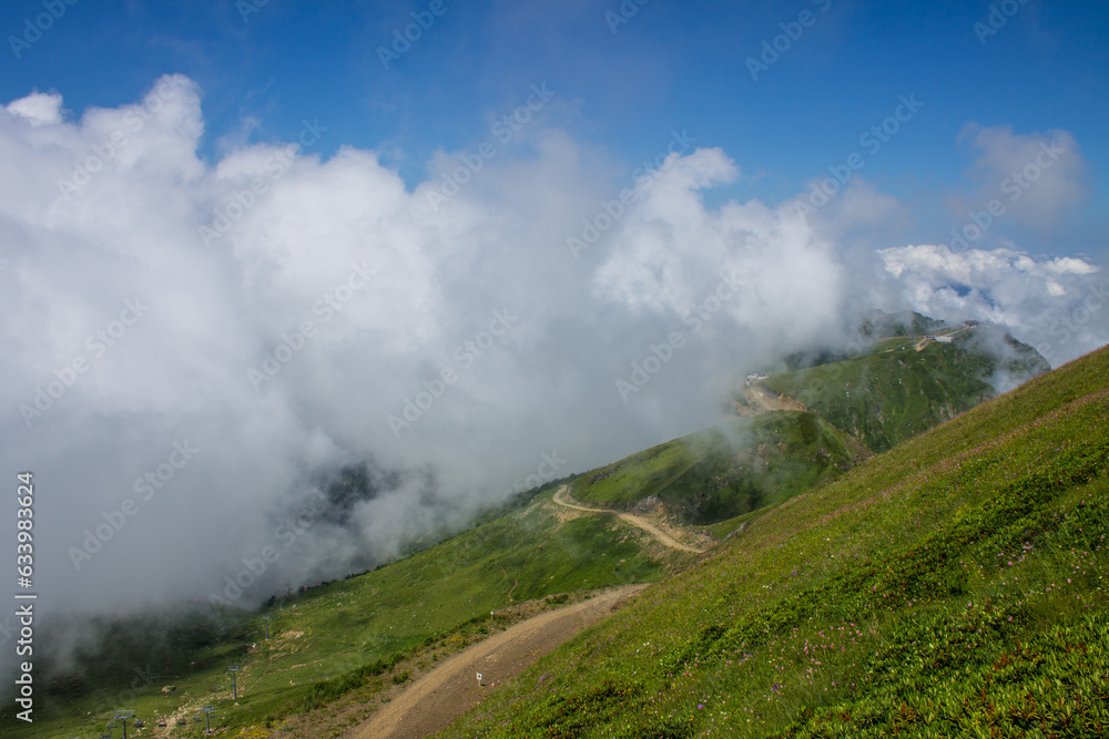 beautiful panoramic landscape - hills with green grass and a walking path among a white big cloud on a red meadow on a summer sunny day and copy space