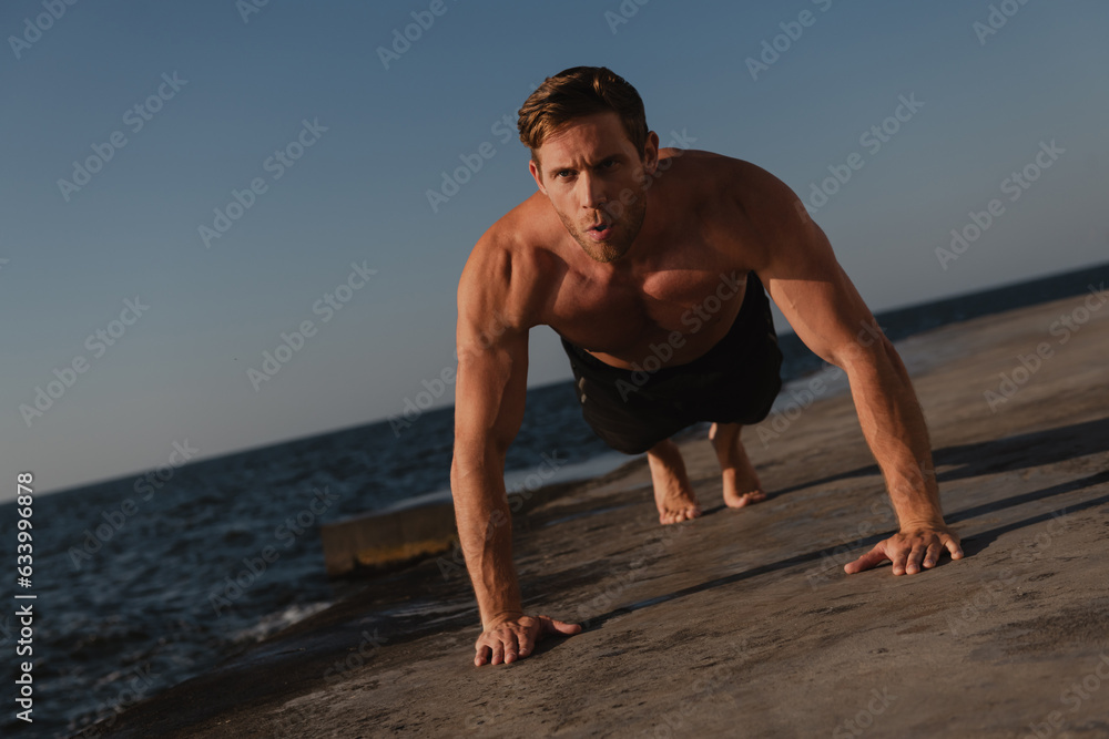 Obraz premium Confident young shirtless man doing push-ups while exercising outdoors with the sea on background