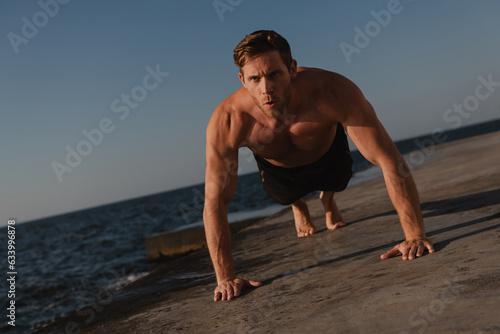 Confident young shirtless man doing push-ups while exercising outdoors with the sea on background