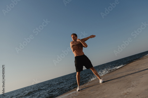 Full length of muscular man doing stretching exercises outdoors with the sea on background