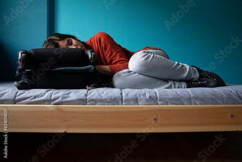 Caucasian woman asleep on a suitcase. Room with a bed. Concepts: dreaming of traveling, traveling, dream, freedom, fantasy, nomad, tired of traveling, Around the World. Blue background wall. (ID: 634014031)