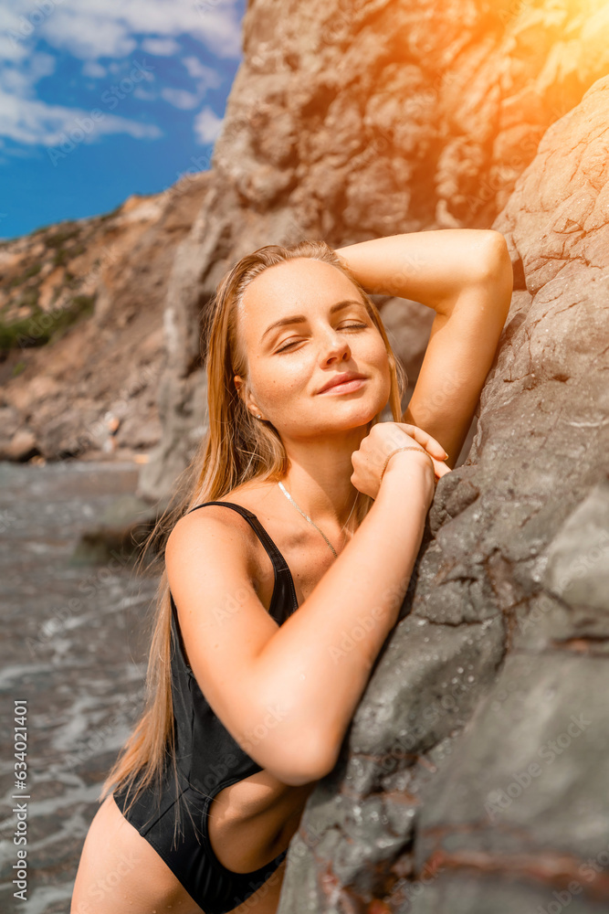 Woman travel sea. Attractive blonde woman in a black swimsuit enjoying the sea air on the seashore around the rocks. Travel and vacation concept.