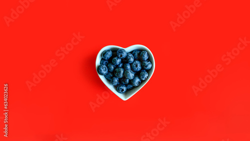 Creative summer layout of blueberries in white heart shaped bowl on vibrant red background. Original blueberry decoration. Minimal summer concept. Welcome summer. Copy space.
