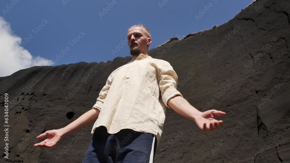 One man in a shirt and with a pigtail on his head stands meditating close to a black rock and blue sky. Calm young blond man meditating with his eyes closed and arms outstretched to the sides One man in a shirt and with a pigtail on his head stands meditating close to a black rock and blue sky. Calm young blond man meditating with his eyes closed and arms outstretched to the sides