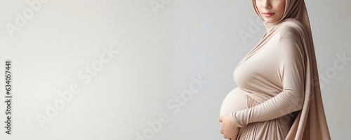 Pregnant muslim woman in hijab embracing her belly over light background with copy space.