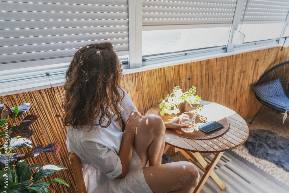 Young caucasian woman in a white shirt sitting on a balcony with roller shutters. Sun protection and heat concept