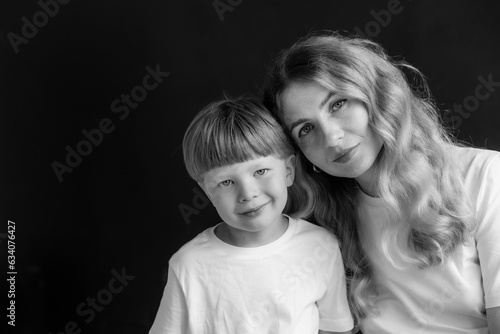 Mom and son at a photo shoot in the studio. The mother and the boy are wearing a white t-shirt. Close-up portrait of mother and son. The background of the photo is black. Black and white photo.