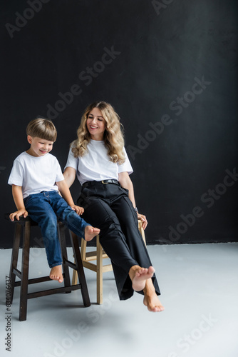 Mom and son at a photo shoot in the studio. Full length portrait. Both of them are wearing a white T-shirt. Mom and son sit on a chair and dangle their legs and smile. The background is black.