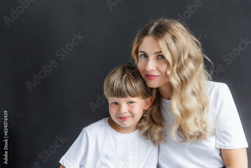 Mom and son at a photo shoot in the studio. The mother and the boy are wearing a white t-shirt. Close-up portrait of mother and son. The background of the photo is black.