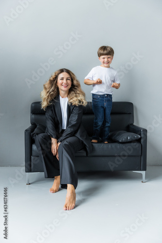 Mom and son at a photo shoot in the studio. Mom is sitting on the sofa. The boy next to him is standing on the couch and smiling. Mom is wearing a black pantsuit. The boy is wearing a white T-shirt.
