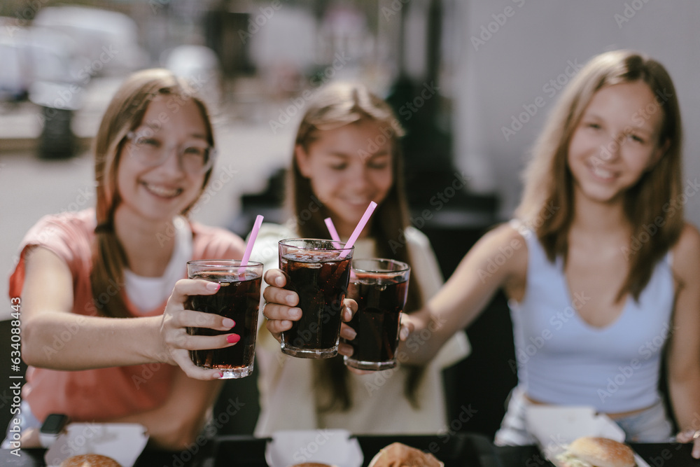 Three joyful smiling teenage girls (14-15 years old) sitting at a cafe outdoors, enjoying cola fresh drinks. Young ladies clinking coke glasses, ready to drink them with pleasure. Focus on glasses