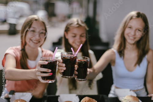 Three joyful smiling teenage girls (14-15 years old) sitting at a cafe outdoors, enjoying cola fresh drinks. Young ladies clinking coke glasses, ready to drink them with pleasure. Focus on glasses