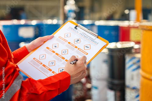 Action of safety officer is using a pen to checking on the hazadous material symbol label form with the chemical barrel as blurred background. Safety industrial working scene concept. Selective focus. photo