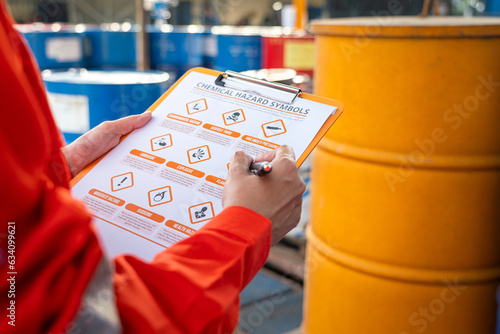 Action of safety officer is using a pen to checking on the hazadous material symbol label form with the chemical barrel as blurred background. Safety industrial working scene concept. Selective focus. photo