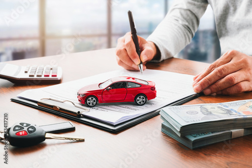 Car loan or buying a new car concept. Red car on clipboard and car key on table with a man signing on lease or document  form in background. Vehicle or automotive business finance concept. 