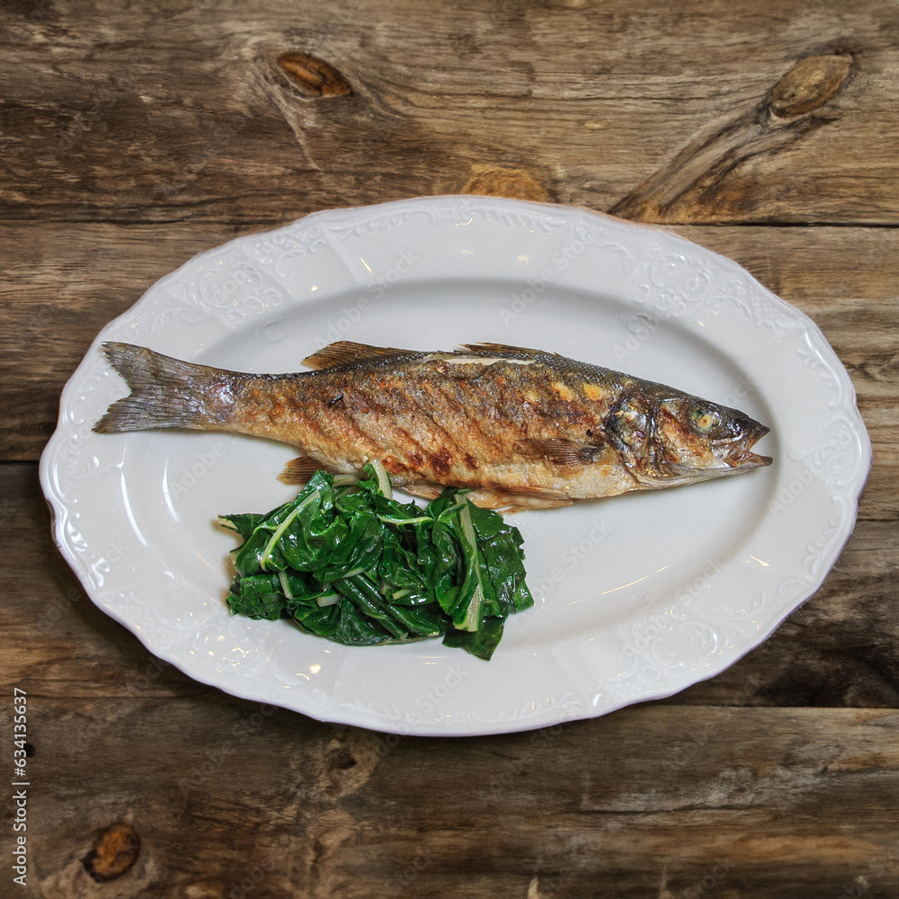 Grilled fish Seabass with spinach garnish served on a white plate on a wooden rustic table Grilled fish Seabass with spinach garnish served on a white plate on a wooden rustic table