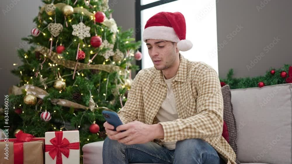 Young hispanic man wearing christmas hat using smartphone upset at home Young hispanic man wearing christmas hat using smartphone upset at home