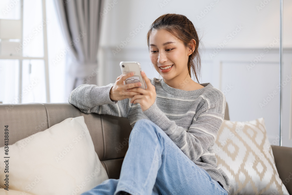 Happy and cheerful woman is using a phone sitting on a sofa.Asian young woman looking mobile phone for shopping online at home. Happy and cheerful woman is using a phone sitting on a sofa.Asian young woman looking mobile phone for shopping online at home.