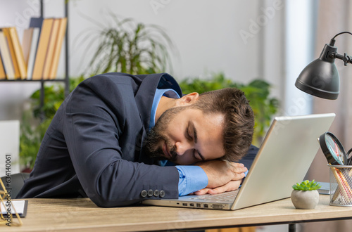 Bored sleepy Caucasian businessman worker working on laptop computer, leaning on hand falling asleep at home office. Exhausted tired freelancer workaholic young man. Employment, occupation, workless photo