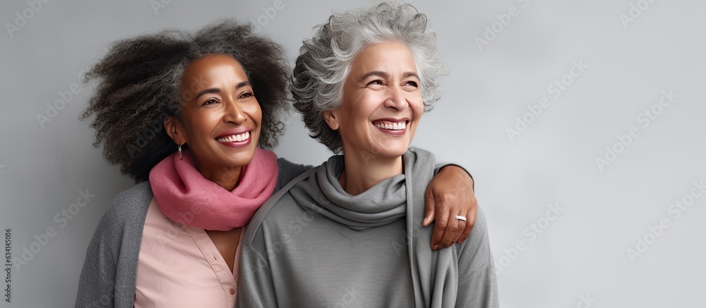 Two joyful middle aged women hugging and laughing standing against a grey wall