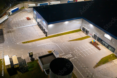 Aerial view of a warehouse of goods at night. Many warehouses and trucks with trailers are in the parking lot, waiting to be loaded for further distribution of goods around the country.