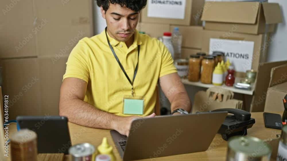 Young latin man volunteer using laptop and touchpad sitting on table at charity center Young latin man volunteer using laptop and touchpad sitting on table at charity center