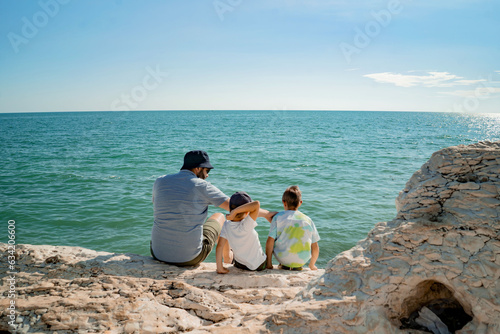 Traveller man and children enjoying beautiful sea view from rocky shore. Father and sons on summer vacation © Yulia Raneva