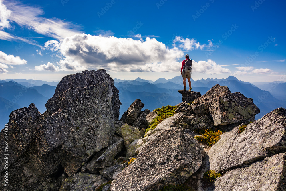 Adventurous athletic female hiker standing on top of a rugged mountain in the Pacific Northwest with jagged mountains in the background.